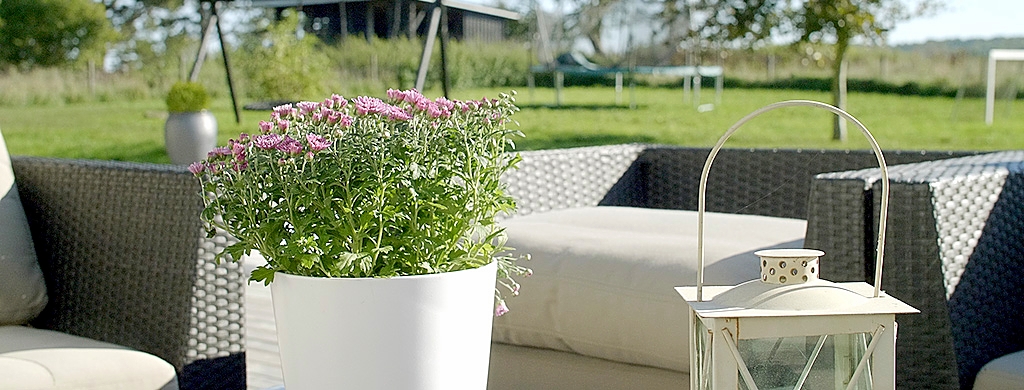 Hvid urtepotte med små lyserøde blomster står på en terrasse foran en lys hynde og et mørkt flettet havemøbelsæt. Til højre står en hvid metallanterne med buet bøjle, og i baggrunden ses en solbeskinnet græsplæne og legeudstyr.