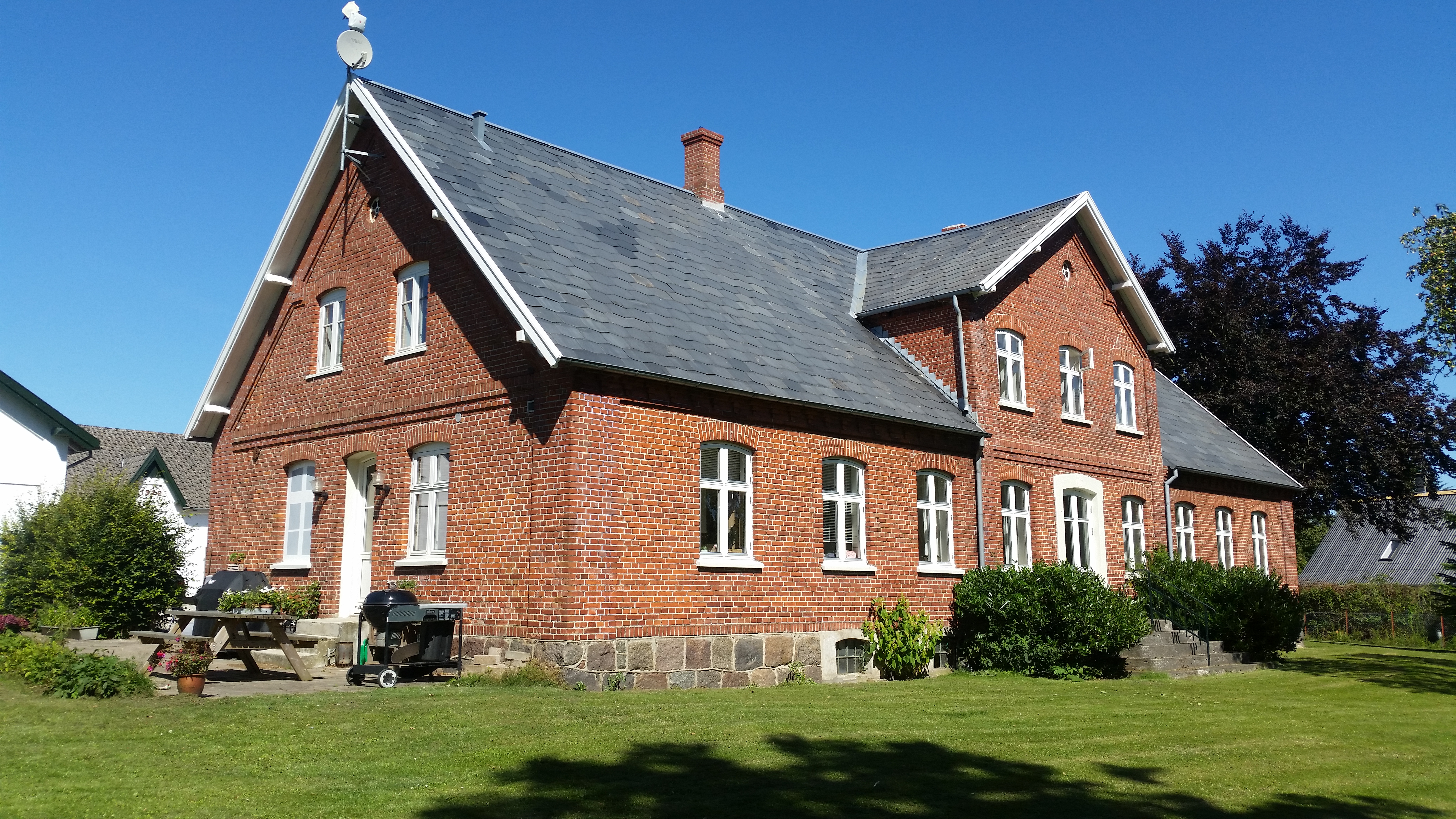 Rødt murstenshus med blågråt skiffertag og hvide vinduesrammer set skråt forfra i en solrig have. Foran huset ligger en velklippet græsplæne, en træ-picnicbænk, to grillapparater og en satellitantenne på tagryggen.