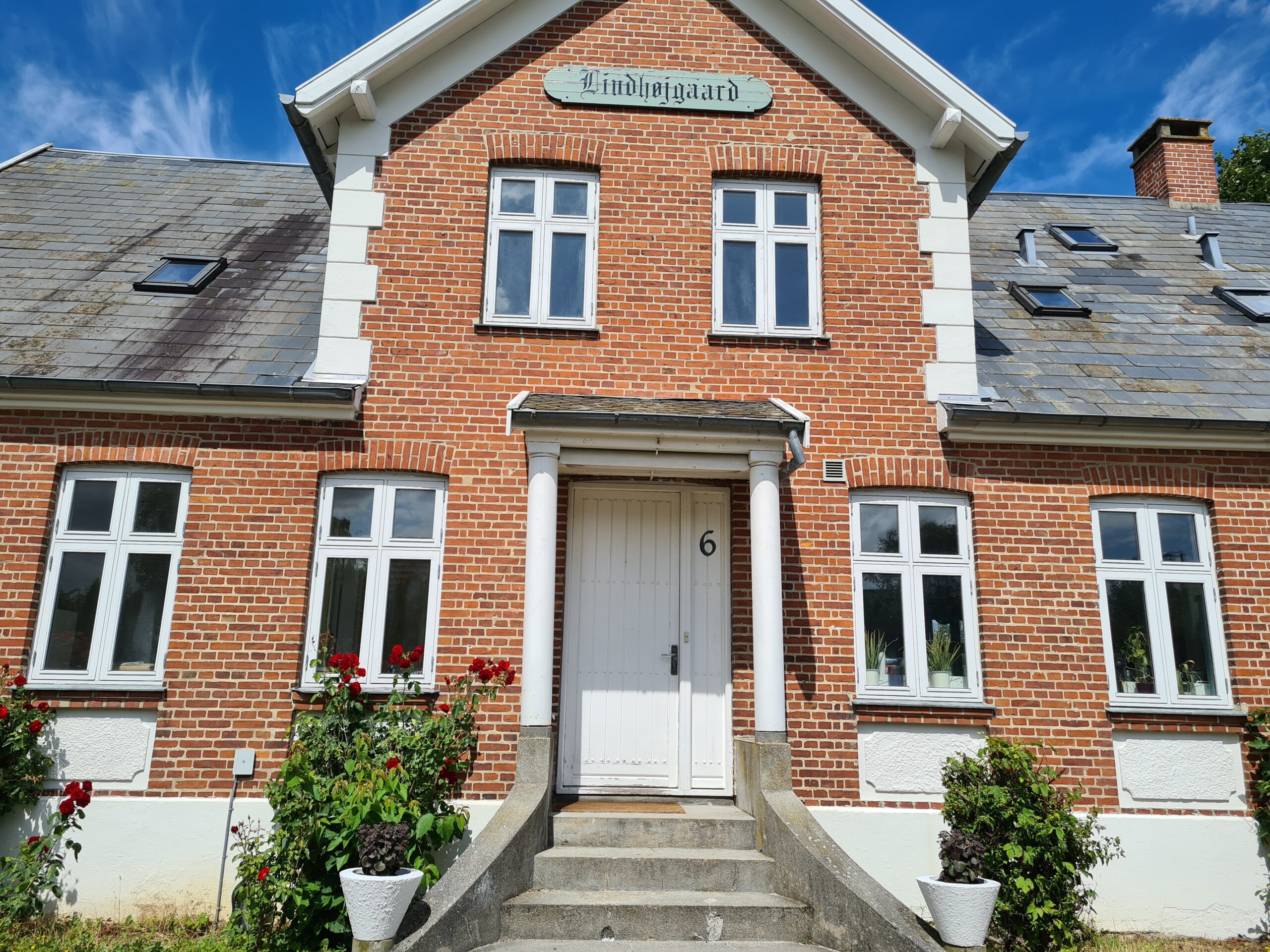 Facaden af et rødt murstenshus med trapper op til en hvid hoveddør mærket med tallet 6 og flankeret af to søjler. Over gavlen ses et skilt med teksten "Lindhøjgaard", og foran huset er blomstrende rosenbuske, krukker samt et skifertag med ovenlysvinduer.