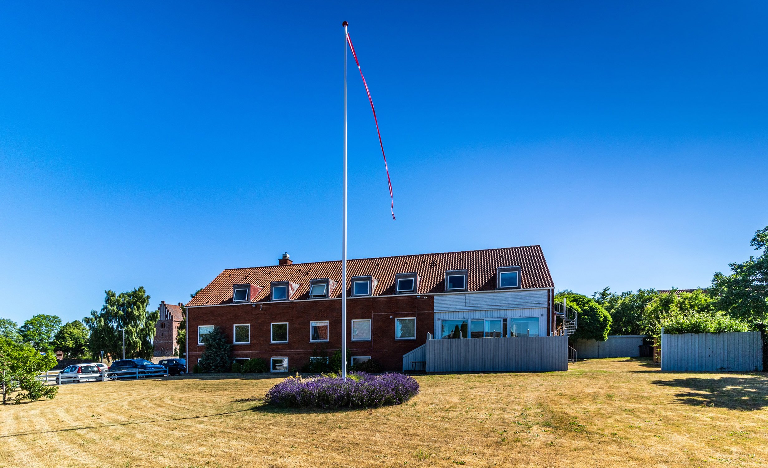 Rødt teglstenshus med rødt tegltag set fra en stor tør græsplæne, midt for en høj flagstang med en lang rød-hvid vimpel der blafrer i vinden; en lav rund lavendelbusk omkranser flagstangen og biler ses parkeret til venstre under klar blå himmel.