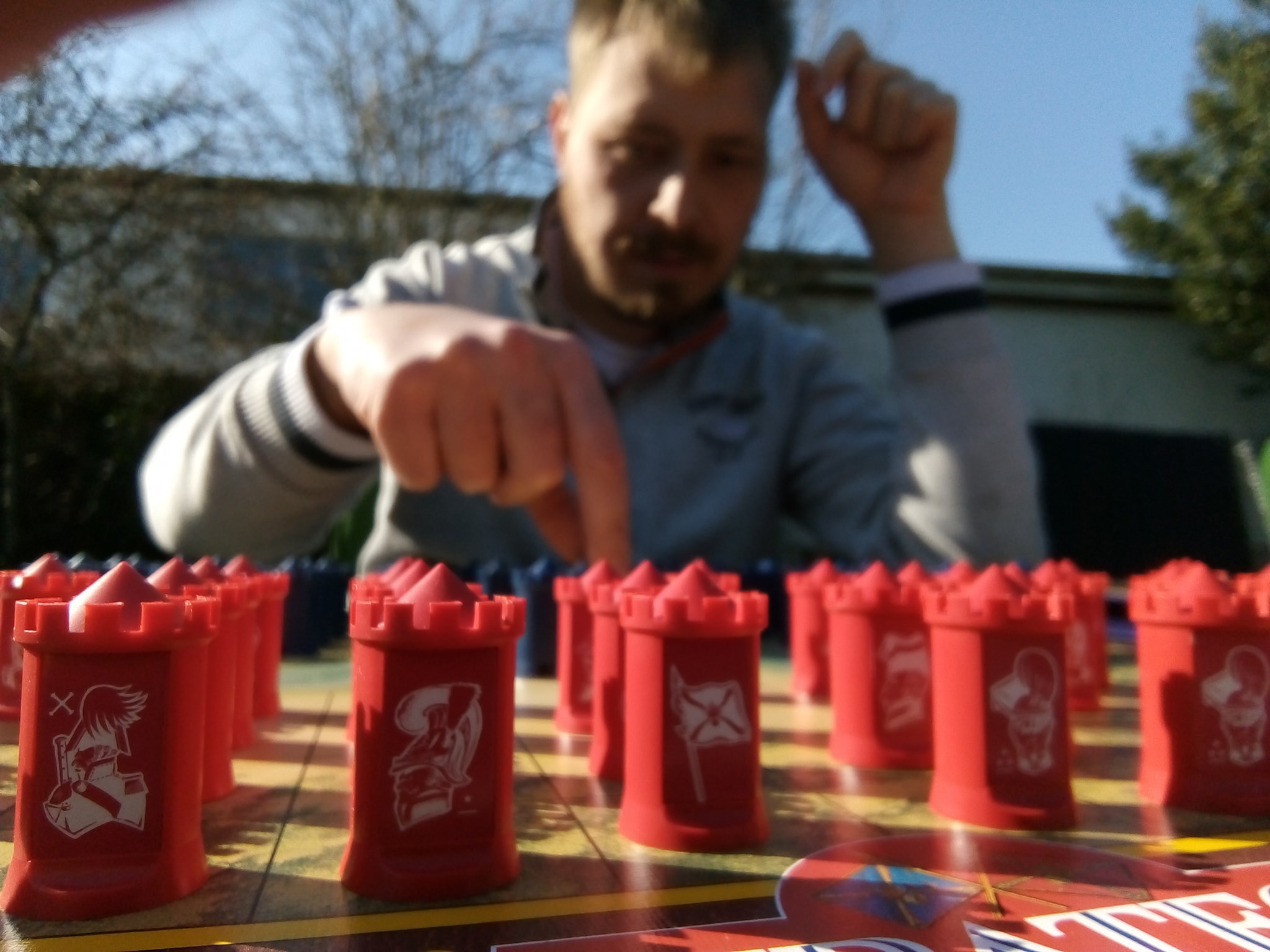 Røde plastik tårnformede brikker på et brætspil i forgrunden, mens en person med sløret ansigt rækker frem og peger på en brik ved et udendørs bord i solskin.