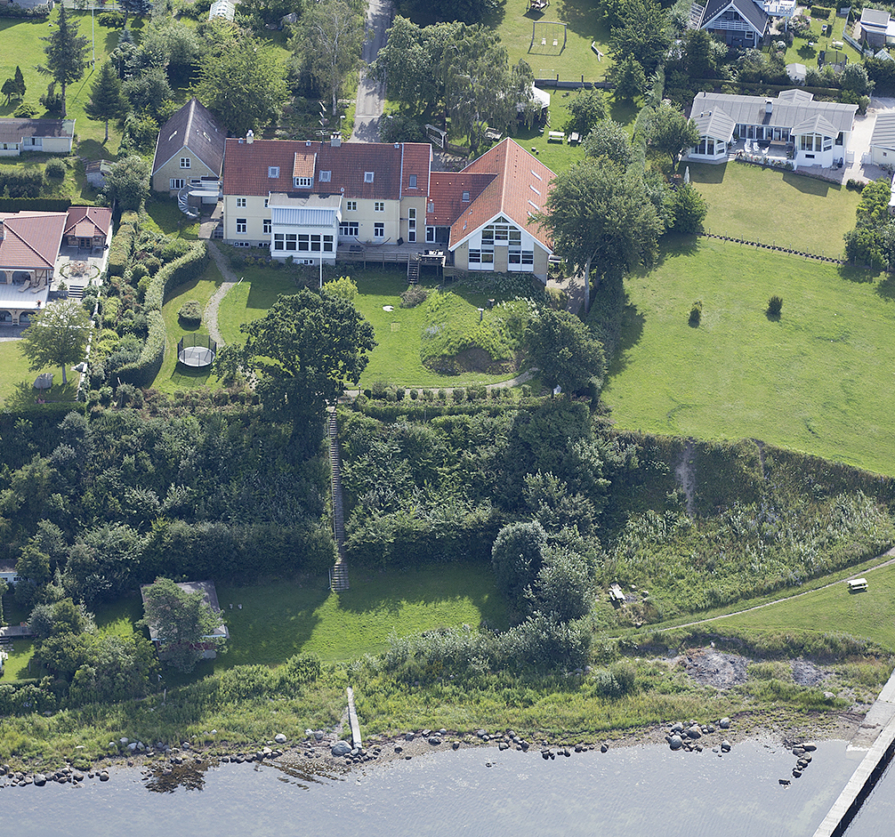 Luftfoto af en stor gul villa med rødt tegltag og glasudestue, omgivet af velplejede plæner, bede og en rund trampolin i haven; nabohuse og åbne græsarealer ses omkring. En trappe gennem buskads fører ned til en smal kyststribe med sten og en lille træbro ud i vandet.