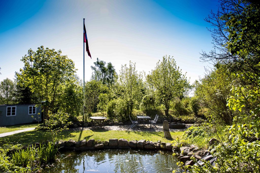 En lille dam med stenrand i forgrunden, foran et græsareal med et udendørs bord og stole omgivet af grønne træer og buske. En høj flagstang med flag står nær midten, og en grå skurbygning ses til venstre under en klar blå himmel.