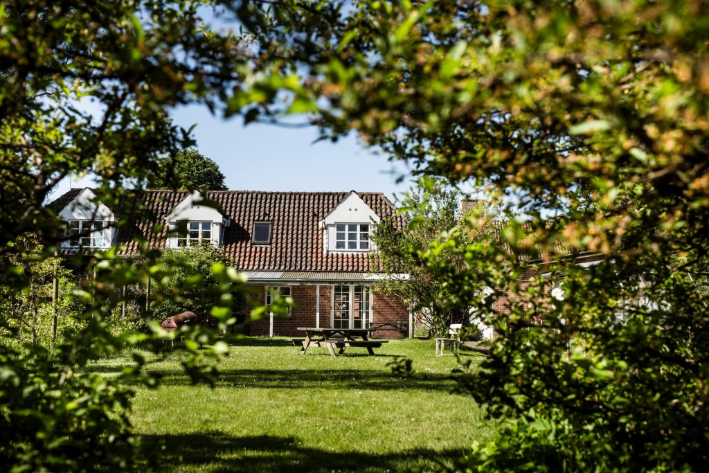 Rødt teglhus med hvide kvistvinduer og rødt tegltag set gennem en ramme af grønne buske og træer. Foran huset en solbeskinnet græsplæne med en træ-picnicbænk og en enkelt hvid stol.