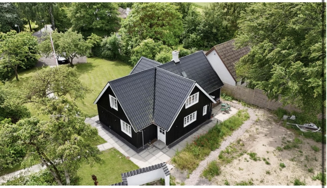 Sort træhus med hvide vinduesrammer og gråt saddeltag set skråt oppefra, placeret på en flisebelagt terrasse. Det er omgivet af frodige træer og græs, med en sandet haveflade og en lille båd ved et hegn samt et nabohus i baggrunden.