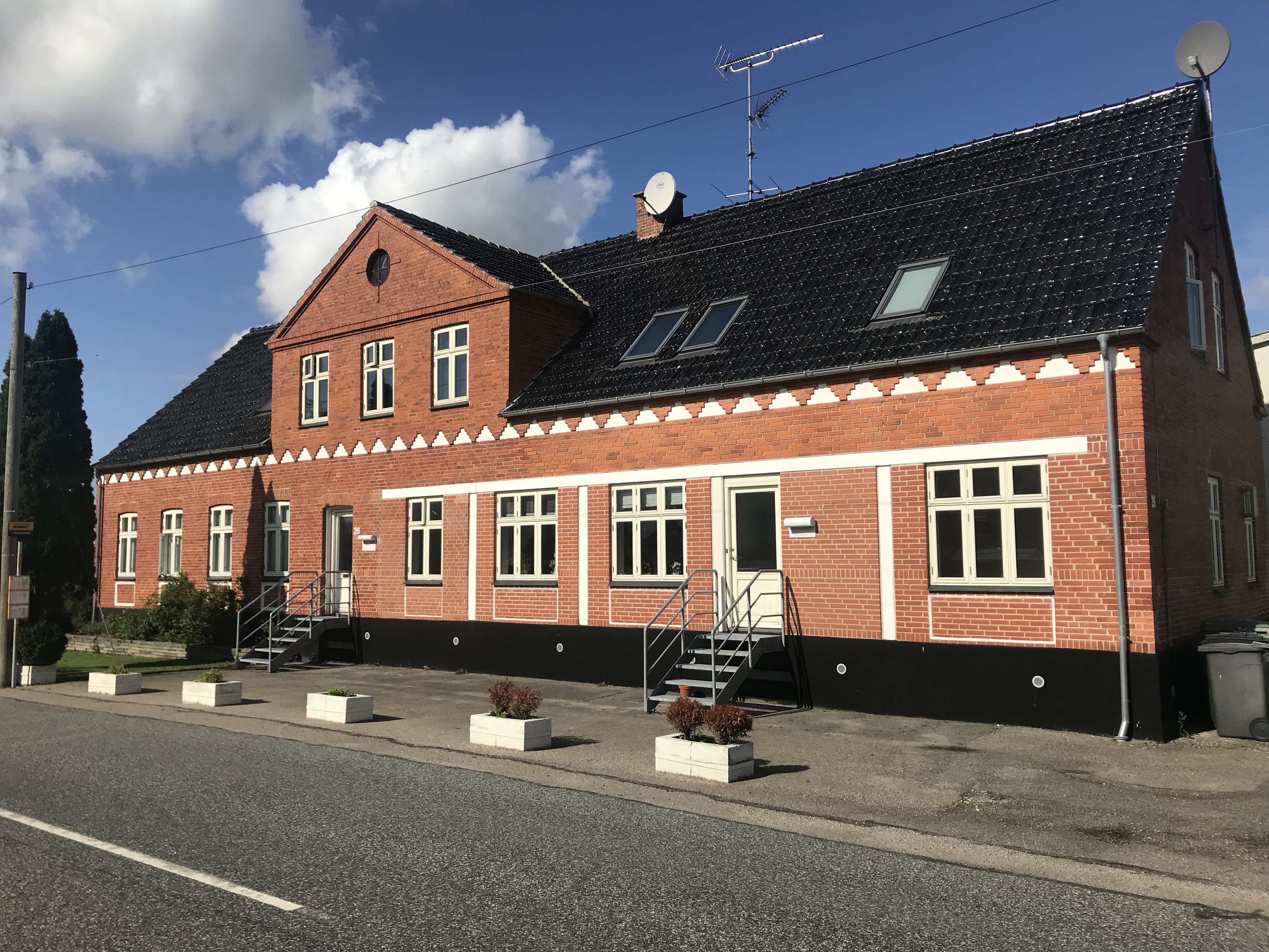 Rødt to-etagers murstenshus med sort saddeltag og hvide vinduesrammer set fra fortovet; to metaltrapper fører op til indgangsdøre, og satellitantenner sidder på taget. Blå himmel med få hvide skyer og fire hvide plantekasser står langs fortovets kant.