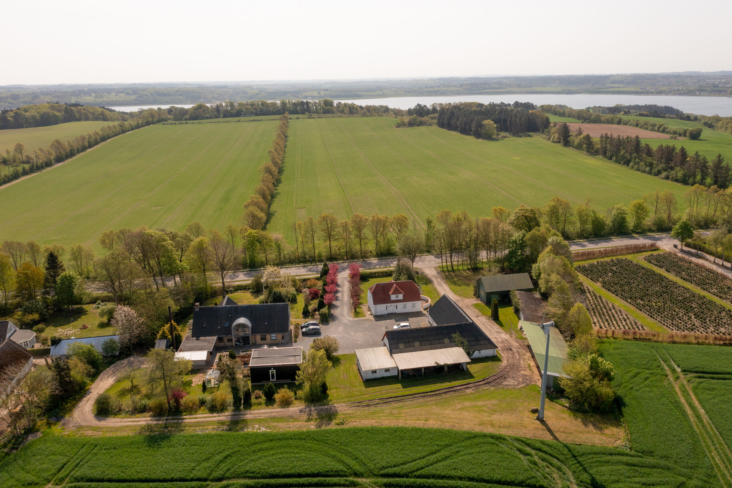 Luftfoto af en landbrugsejendom set ovenfra med flere gårdsbygninger og en gårdsplads. Ejendommen er omgivet af store grønne marker, træalléer langs en vej og en sø i horisonten.