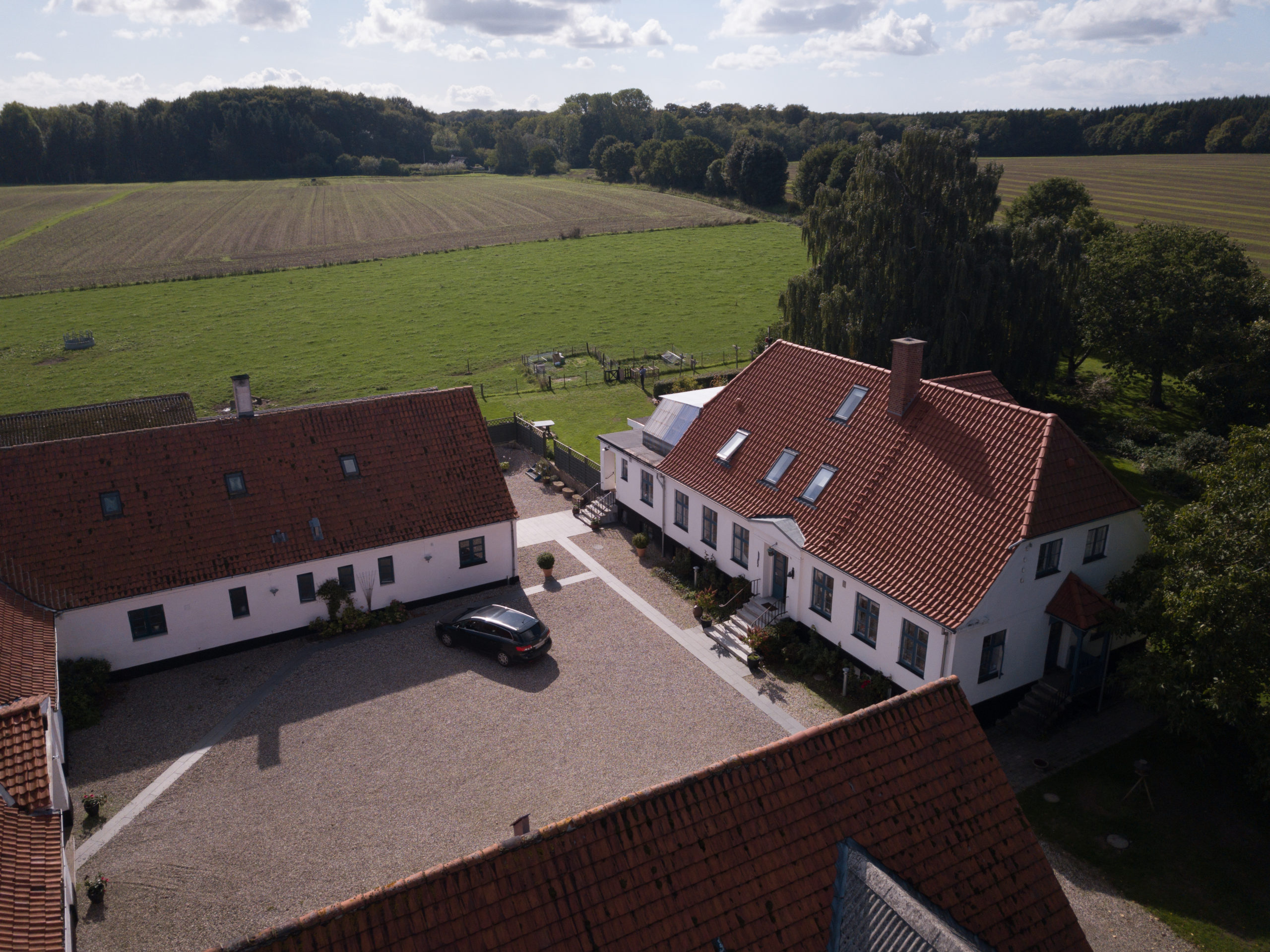 Luftfoto af en gårdsplads med hvide bygninger med røde tegltage og en gruset plads, hvor en sort bil holder parkeret, og et af tagene har solpaneler. Omkring gården ligger grønne marker og træer under en delvist skyet himmel.