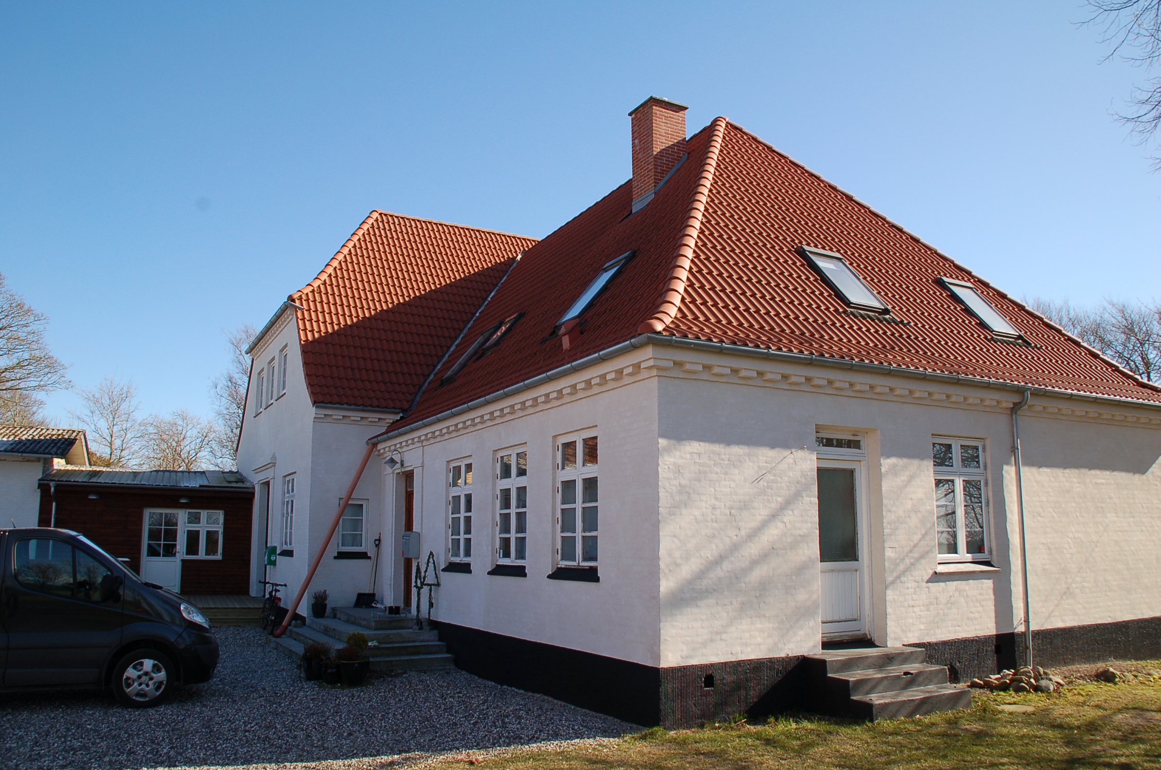 Hvidt murstenshus med markant rødt tegltag, skrå tagvinduer, skorsten og små trapper ved indgangene, set i en grusbelagt gårdsplads. En mørk varevogn holder til venstre og nøgne træer ses i baggrunden under en klar blå himmel.