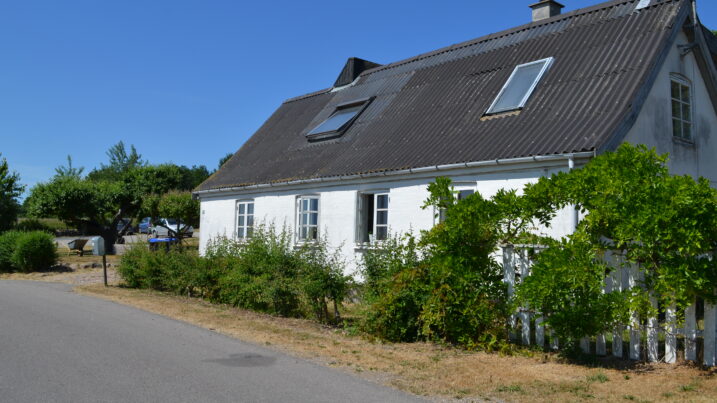 Hvidt étplans hus med mørkt bølgeformet tag og to ovenlysvinduer samt et åbent facadevindue, omgivet af grønne buske og et hvidt stakit under en klar blå himmel. En asfaltvej løber forbi huset i forgrunden.