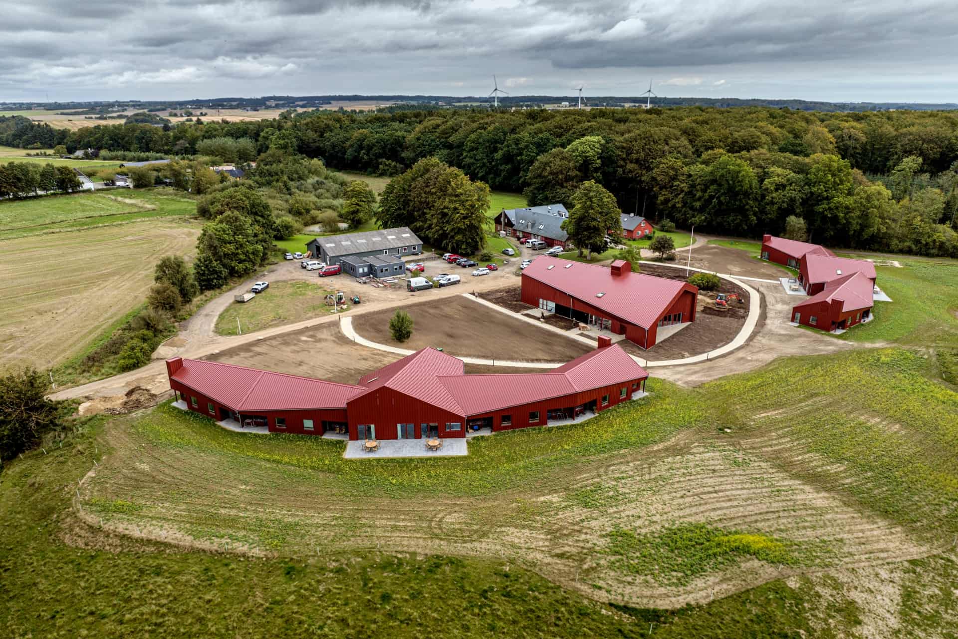 Luftfoto af en nyanlagt klynge røde bygninger med røde tage i en buet planløsning omkring indre veje og gårdsplads på en grøn mark, omgivet af skov og landbrugsjord under en overskyet himmel. Nogle biler er parkeret ved de centrale bygninger, og vindmøller ses langs horisonten.