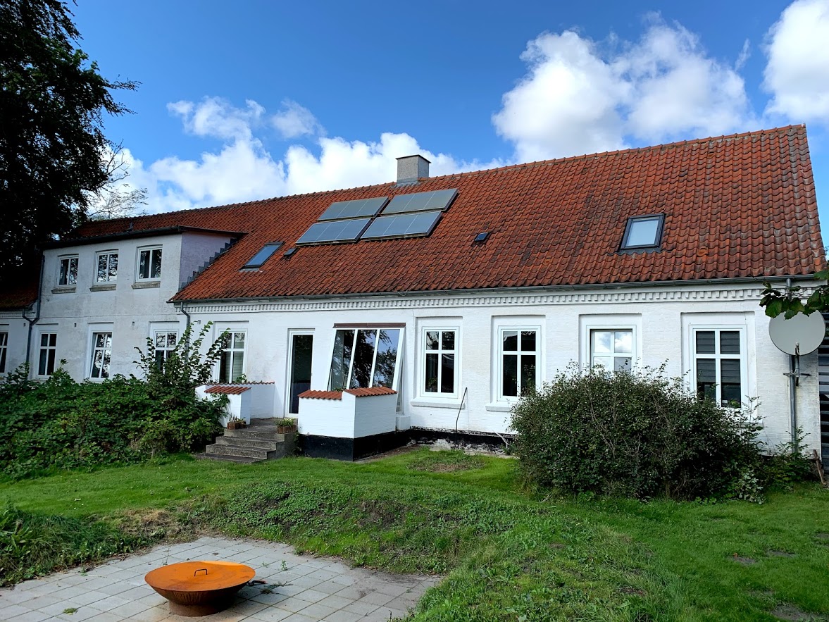 Hvid bygning med rødt tegltag og fire solpaneler på skrå taget, omgivet af buske og en grøn plæne med en orange ildskål placeret på fliser i forgrunden.