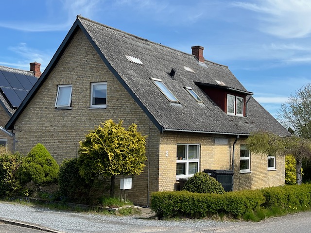 Gulstens parcelhus med stejlt gråt tag og en rød kvist med vinduer, flere ovenlysvinduer og en skorsten, omgivet af lav hæk og buske langs en grusvej under blå himmel; husnummeret "8" er synligt på husmuren.