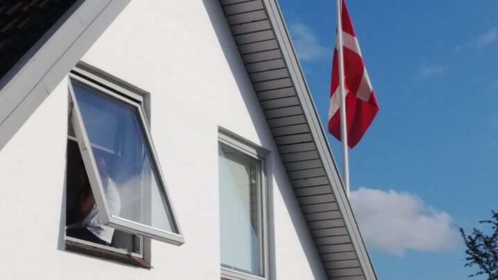 Hvid husgavl med to vinduer, hvor venstre vindue står på klem og en person sidder og kigger ud af åbningen. Dannebrog blafrer på en flagstang ved tagkanten mod en klar blå himmel med enkelte skyer.