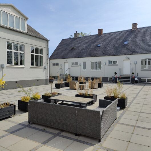 Grå flettet havemøbelgruppe placeret omkring et lavt bord på en stor flisebelagt gårdsplads, omgivet af sorte rektangulære plantekasser med små træer og høje græsser. To lave hvidmalede bygninger med mange vinduer, udvendige trapper og parkerede cykler ses i baggrunden under blå himmel.