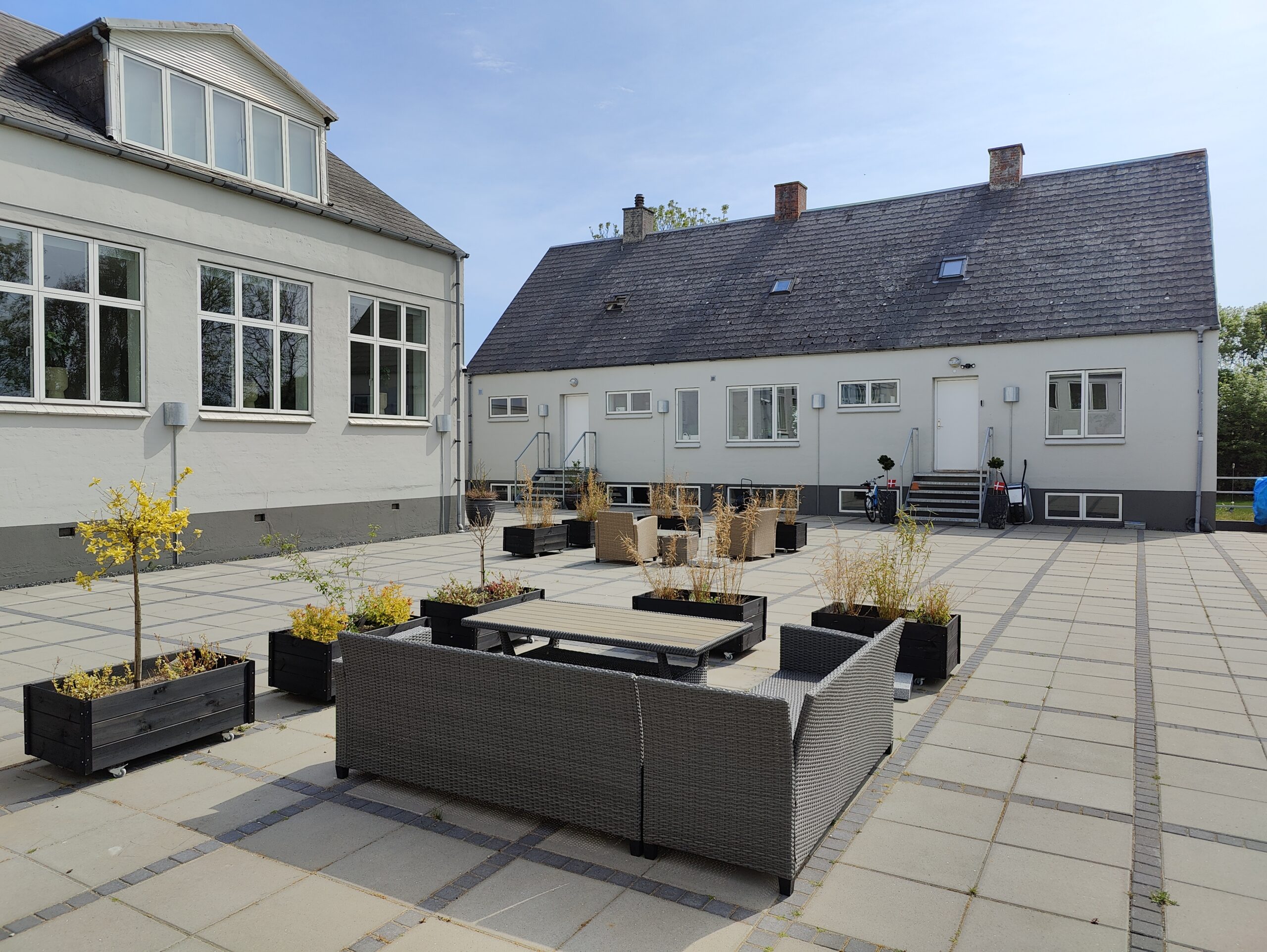 Grå flettet havemøbelgruppe placeret omkring et lavt bord på en stor flisebelagt gårdsplads, omgivet af sorte rektangulære plantekasser med små træer og høje græsser. To lave hvidmalede bygninger med mange vinduer, udvendige trapper og parkerede cykler ses i baggrunden under blå himmel.