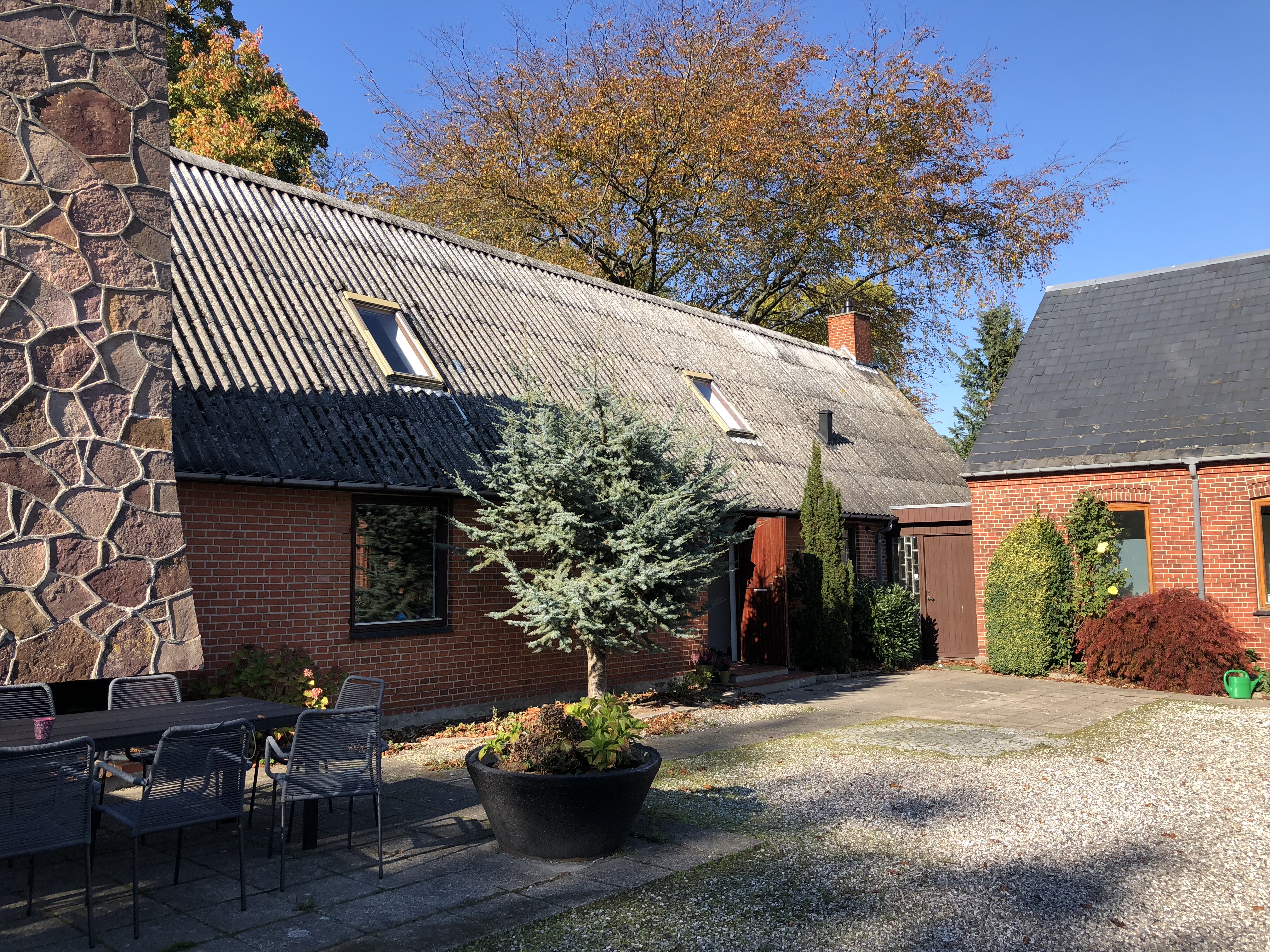 Gårdsplads med en rød murstensbygning og gråt, skråt bølgeeternittag med to skråvinduer og en høj stenvæg til venstre; midt i forgrunden står en stor sort plantekrukke med en lille nåletræsbusk foran et sort metal spisebord med stole. I baggrunden ses efterårsfarvede træer og en klar blå himmel.