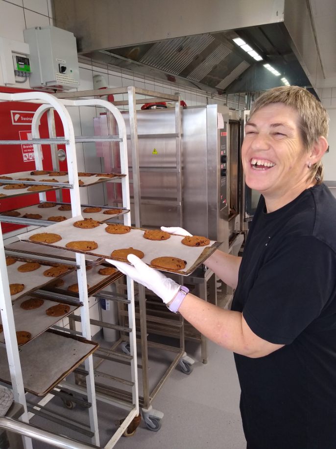 En person med sløret ansigt og hvide handsker holder en bageplade med nystegte chokoladekookies og sætter den ind på et rullestativ i et professionelt køkken foran en stor ovn.
