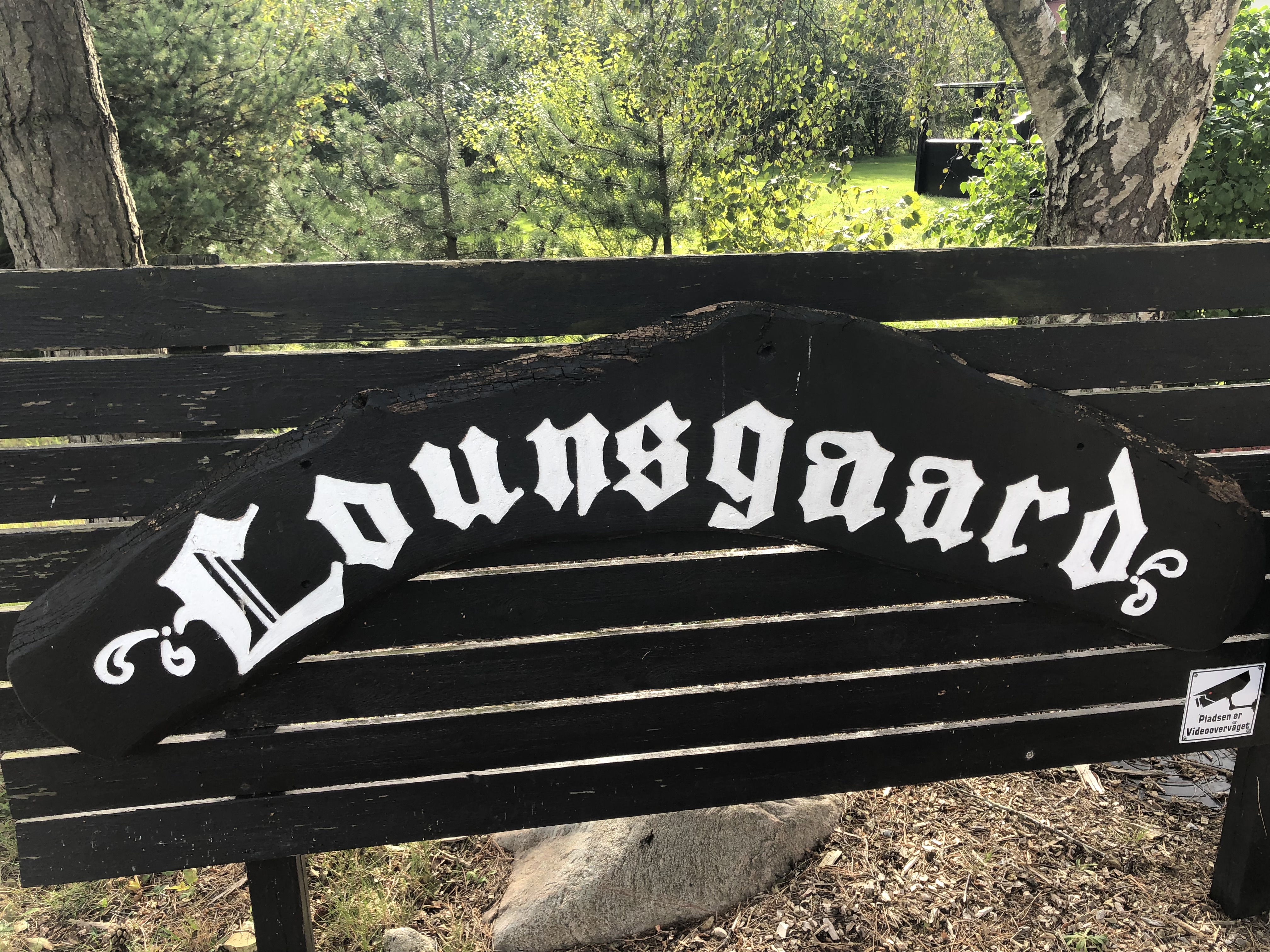 Sort træskilt med hvid gotisk tekst "Lounsgaard" monteret på en sort, lamel-agtig træbænk foran birketræer og græs; nederst til højre sidder et klistermærke med et kameraikon og teksten "Pladsen er Videoovervåget".