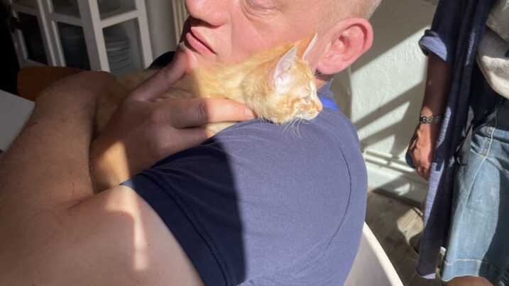 Mand med kortklippet hår holder en lille lysebrun hund tæt ind til sig, mens han sidder på en hvid stol ved et bord i et solbeskinnet rum; en kvinde står til højre i denimnederdel og sandaler.