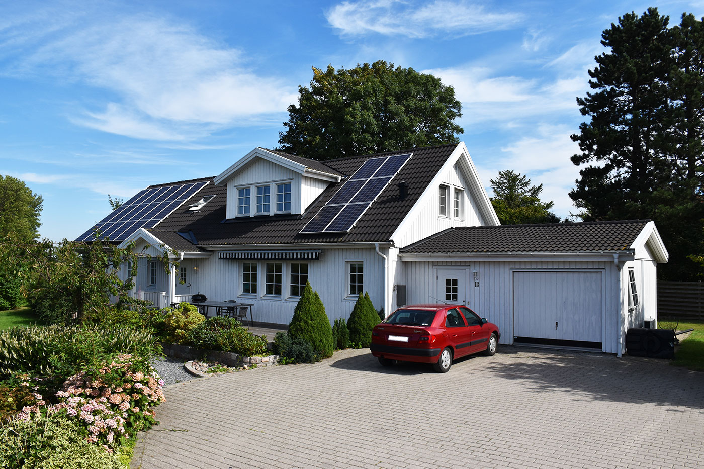 Hvid trævilla med mørkt tag dækket af solcellepaneler og en velholdt have, mens en rød bil står parkeret på flisebelagt indkørsel foran en lukket garage under en blå himmel.