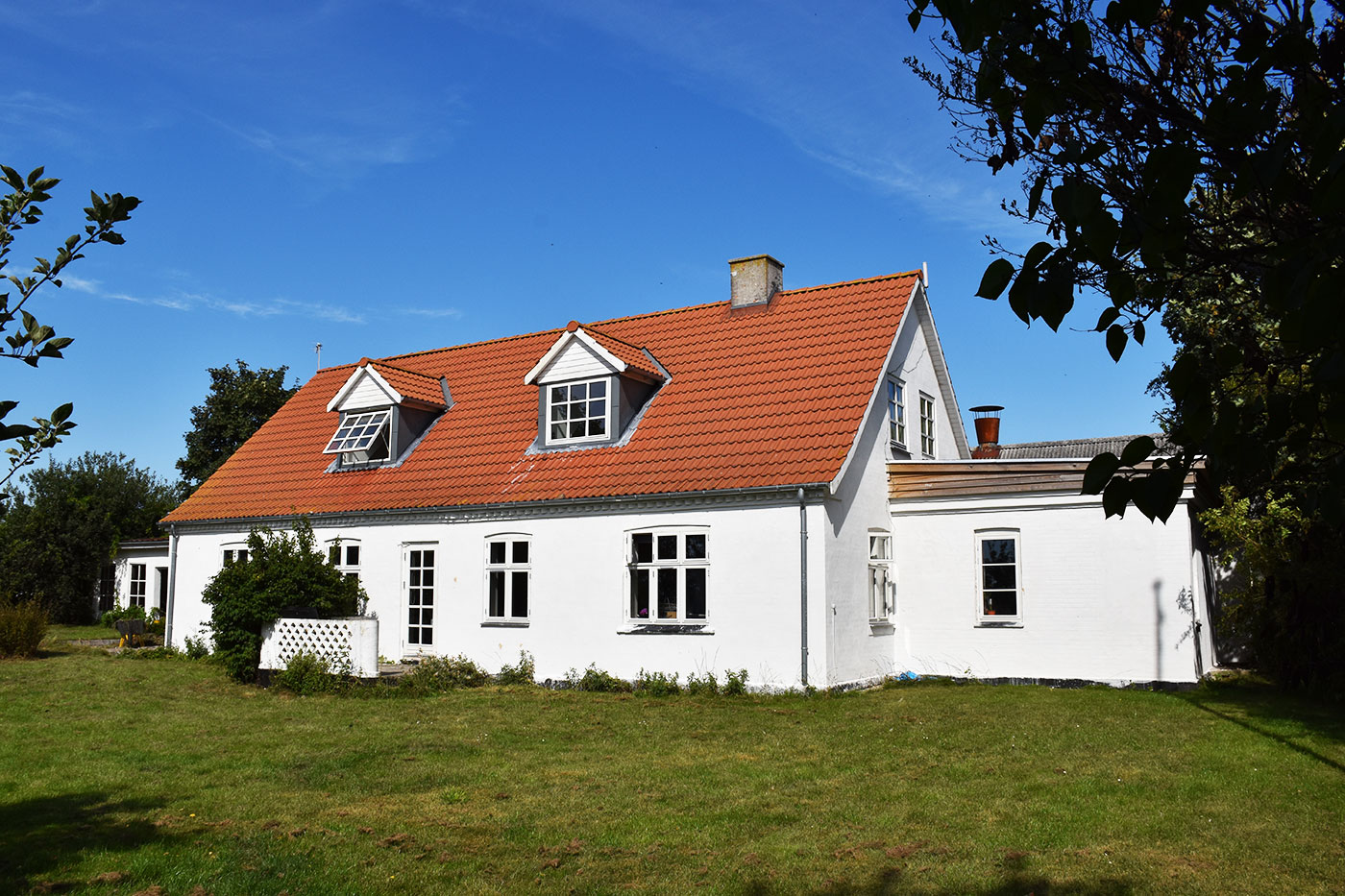 Hvidt hus med rødt tegltag og to kvistvinduer, hvor det ene står åbent, placeret på en rummelig grøn græsplæne under en klar blå himmel. Træer og buske rammer huset ind i for- og baggrunden.