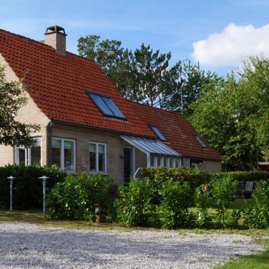 Lys murstensvilla med rødt tegltag og tagvinduer set fra forhaven; foran huset ligger en grusindkørsel og et æbletræ med lave hække og græsplæne, og til højre ses et lille drivhus under en blå himmel.