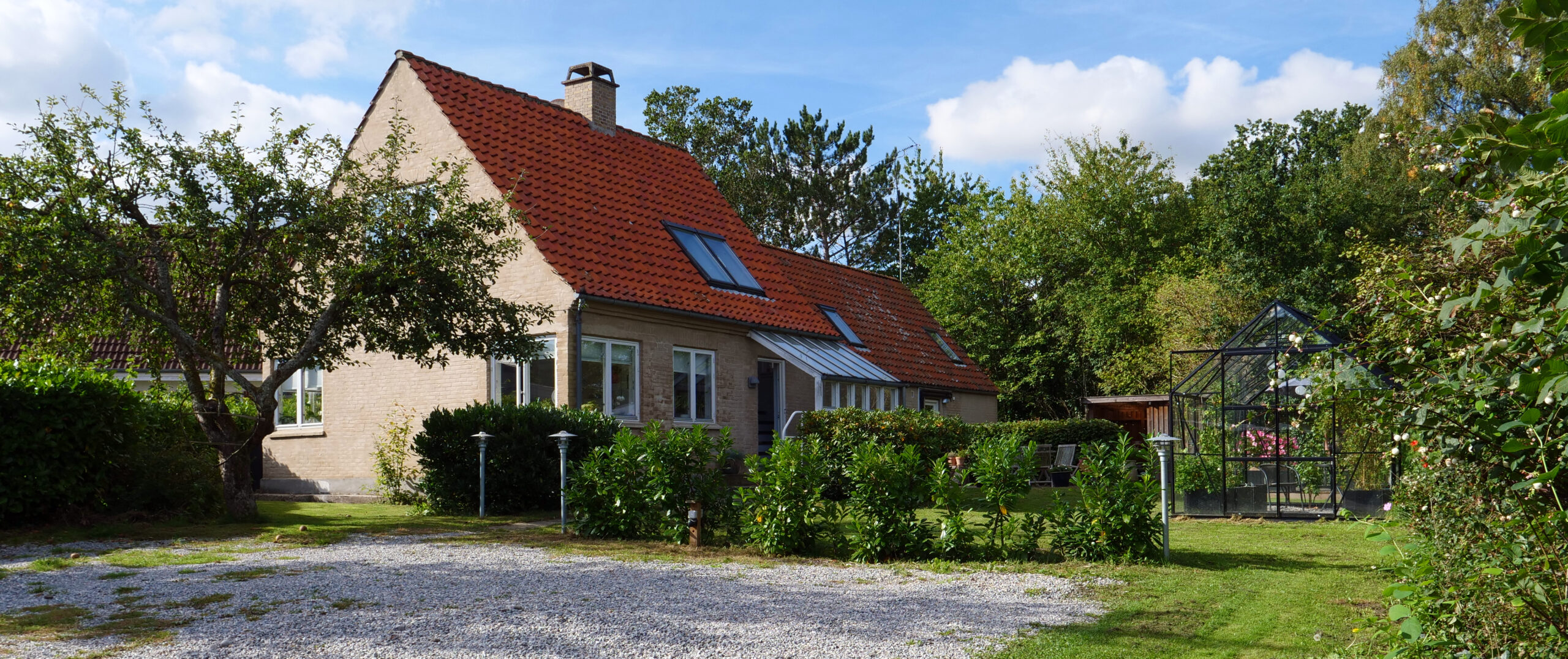 Lys murstensvilla med rødt tegltag og tagvinduer set fra forhaven; foran huset ligger en grusindkørsel og et æbletræ med lave hække og græsplæne, og til højre ses et lille drivhus under en blå himmel.