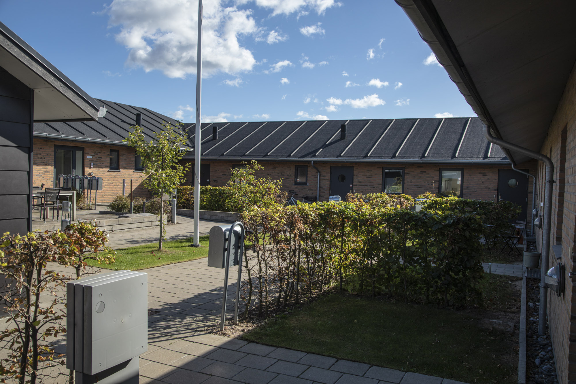 Gårdrum mellem lave rækkehuse i rød mursten med mørkt tag, hvor en flisebelagt sti og trimmede hække fører forbi små træer og en flagstang under en blå himmel. I forgrunden ses en grå elboks og flere postkasser langs stien.