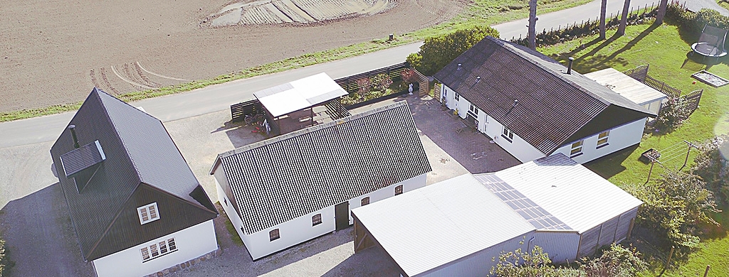 Luftfoto af en landejendom med flere hvide bygninger med mørke tage samlet omkring en gårdsplads, et lille carport‑skur ved vejen, solpaneler på et tag og marker samt en græsplæne med trampolin i baggrunden.