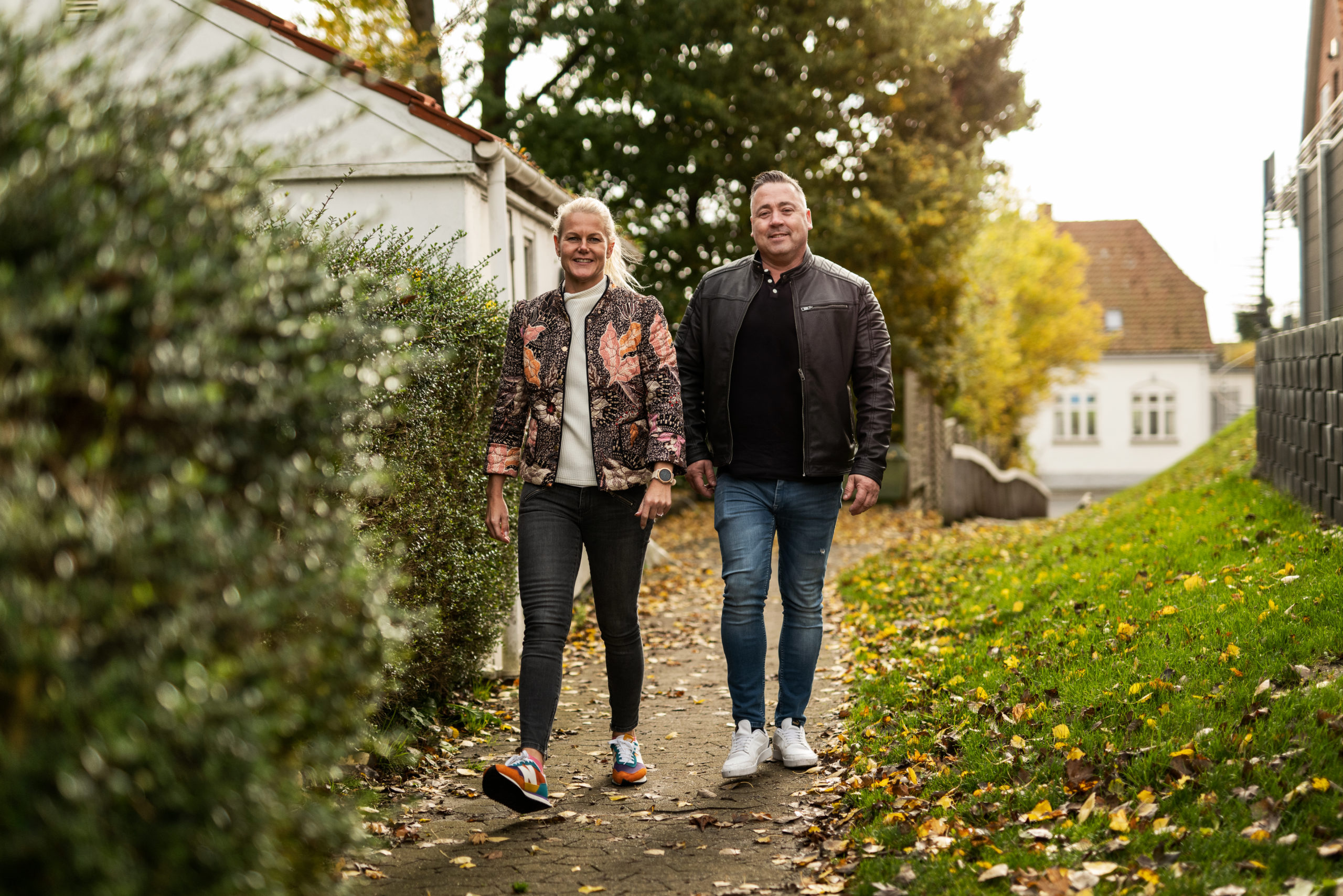 To voksne personer går side om side ned ad en smal fortovssti dækket af efterårsløv i et boligområde; kvinden til venstre bærer en mønstret jakke og farverige sneakers, manden til højre en sort læderjakke og hvide sko.