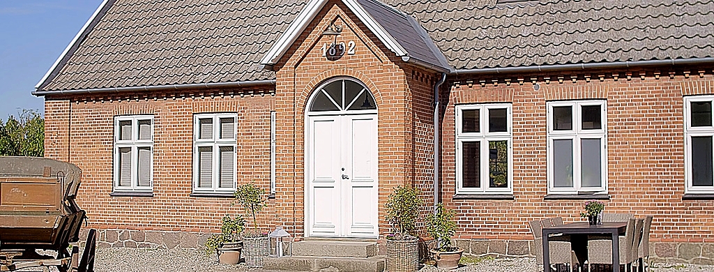 Rød teglstensbygning med en hvid dobbeltdør i midten under en lille gavl mærket u00221892u0022. Symmetriske hvide vinduer flankerer døren, krukker med planter står ved trappen, og et lille udendørsbord med stole er til højre, mens en gammel vogn ses delvist til venstre på gruspladsen.