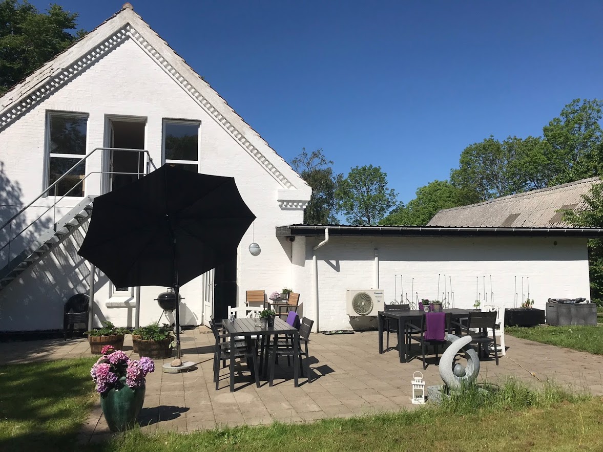 Hvidt to-etagers hus med skrånende tag og en stenbelagt terrasse foran; på terrassen står flere sorte havemøbler og borde, et stort åbent sort parasol, en grill, krukker med lyserøde blomster og en rund dekorationsskulptur, omgivet af grøn græsplæne og træer under en klar blå himmel.