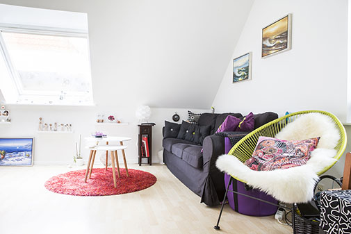 Loftstue i en kvist med skråt tagvindue, lyst trægulv og to små hvide runde borde placeret på et rundt rødt tæppe. Et mørkegråt sofa med flere lilla puder står mod væggen under to malerier, og en limegrøn flettet stol med hvidt fåreskind og mønstret pude står forrest.