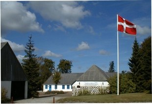 Hvid lav bygning med blå vindueslåger står på en grøn plæne omgivet af træer, mens et rødt-hvidt dansk flag blafrer i vinden på en høj flagstang under en blå himmel med få skyer.