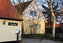 Gult to-etagers hus ved siden af en mindre bygning med et sort skilt mærket "Gården", omgivet af nøgne træer og en gruset indkørsel.