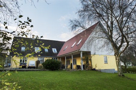 Gult to-etagers hus med rødt skråtag og hvid gavl og en venstre fløj med mørkt tag og tagvinduer, placeret på en velplejet grøn græsplæne med birketræer i forgrunden under en let skyet himmel.