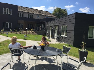 To kvinder sidder og samtaler ved et rundt metalbord på en flisebelagt terrasse; på bordet står en potteblomst og nogle kopper. Terrassen ligger foran moderne lavere bygninger med mørke træfacader, græsplæner og bede under en solrig himmel.