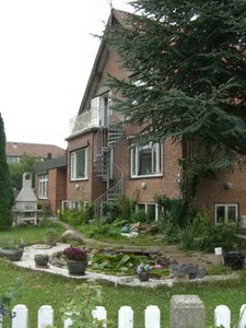 Et rødt murstenshus med en udvendig metal-spiraltrappe der fører op til en lille balkon; foran huset ligger en have med en dekorativ dam omgivet af sten og blomsterpots, en hvid stakit i forgrunden og et stort nåletræ ved siden af huset.