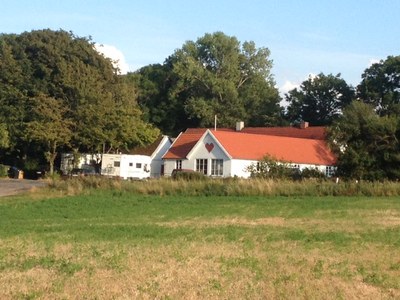 Hvid gårdbygning med rødt tegltag og et stort rødt malet hjerte på gavlen står i en åben græsmark omgivet af træer under en blå himmel.