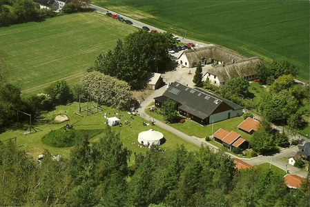 Luftfoto af en landlig gård/fritidscenter med flere bygninger og et åbent græsareal, hvor to hvide telte er opstillet og små grupper af mennesker opholder sig; omgivet af træer og marker med en vej og parkerede biler langs billedets øverste kant.