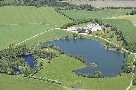 Luftfoto af en gård med bygninger og adgangsvej ved en stor, let hjerteformet sø omgivet af grønne marker; en mindre dam ses til venstre, og træer samt hegn afgrænser landskabet.