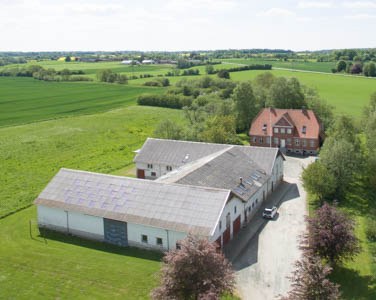 Luftfoto af en landbrugsejendom med et rødt hovedhus bagved og flere hvide udbygninger med grå tage — det nærmeste tag har solceller; omgivet af grønne marker, træer og en grusvej med en hvid bil parkeret.