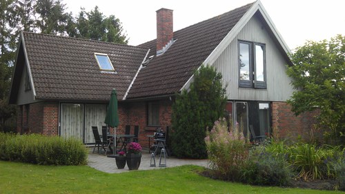 Et fritliggende enfamiliehus i to etager med rød murstensfacade og lysmalet træbeklædning, skråt tag med skorsten og ovenlysvindue. Foran huset en flisebelagt terrasse med havebord, stole, sammenklappet grøn parasol og blomsterkrukker, omgivet af græsplæne og buske.