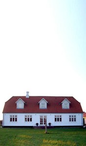 Et hvidt, toetagers hus med rødt tegltag og tre kvistvinduer på facaden står midt i en velklippet grøn plæne under en klar, lyseblå himmel.