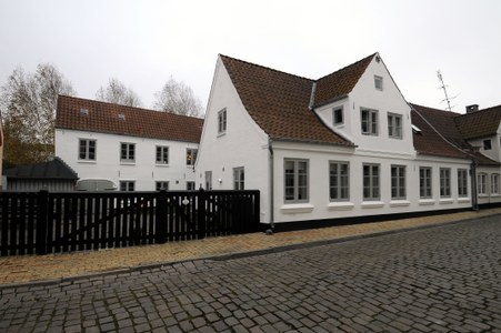 Hvidt to-etagers hus med rødt tegltag og mange hvide sprossevinduer, som står langs en brostensbelagt gade foran et sort træhegn under en grå overskyet himmel.