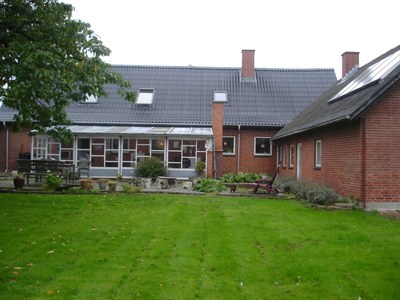 Rød murstensrækkehus med glasveranda og flere skorstene set bag en stor, velplejet græsplæne; terrasse med plantekasser og havemøbler foran, et træ til venstre og skrånende tage med tagvinduer.