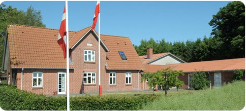 Rødt murstenshus med rødt tegltag og hvide vinduesrammer, to danske flag på flagstænger i forgrunden; huset er omgivet af lave buske, græs og træer under en klar blå himmel.
