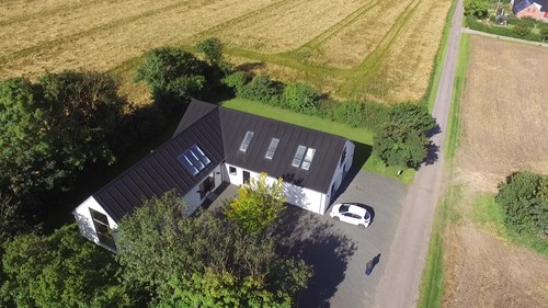 Luftfoto af et L-formet hvidt hus med mørkt tag og flere ovenlysvinduer, omgivet af træer og kornmarker. En hvid bil er parkeret ved indkørslen til huset langs en smal vej, og en person ses som en skygge ved indkørslen.