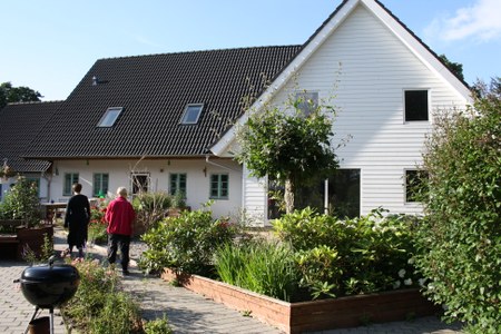 Hvidt to-etagers hus med mørkt tag og en flisebelagt sti gennem en frodig forhave; to personer, én i sort og én i rød jakke, går væk mod huset, mens en rund sort grill står i forgrunden til venstre.